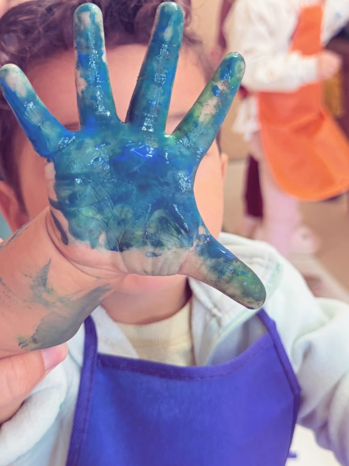 Kinderhand voller blauer Farbe, das Kind trägt eine Malschürze